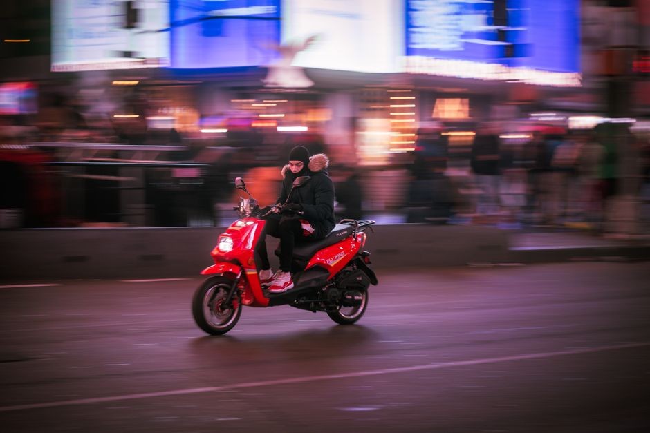 Seseorang mengendarai skuter merah melalui jalan kota yang ramai di malam hari.