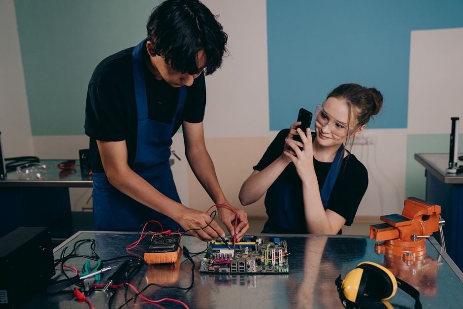 Dua orang dewasa muda menguji motherboard di laboratorium teknologi, merekam prosesnya dengan smartphone.