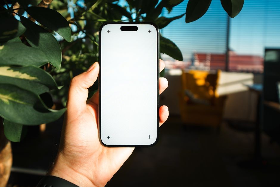 Sebuah tangan dewasa memegang smartphone dengan layar kosong di ruangan yang nyaman dan terang, sempurna untuk mockup digital.