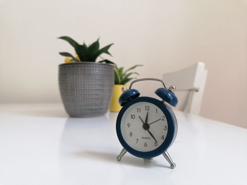 Jam alarm biru klasik di atas meja putih dengan tanaman pot, cocok untuk tema dekorasi modern.