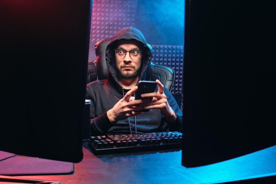 Seorang pria berbaju hoodie menggunakan smartphone, dikelilingi oleh peralatan teknologi di ruangan yang redup.