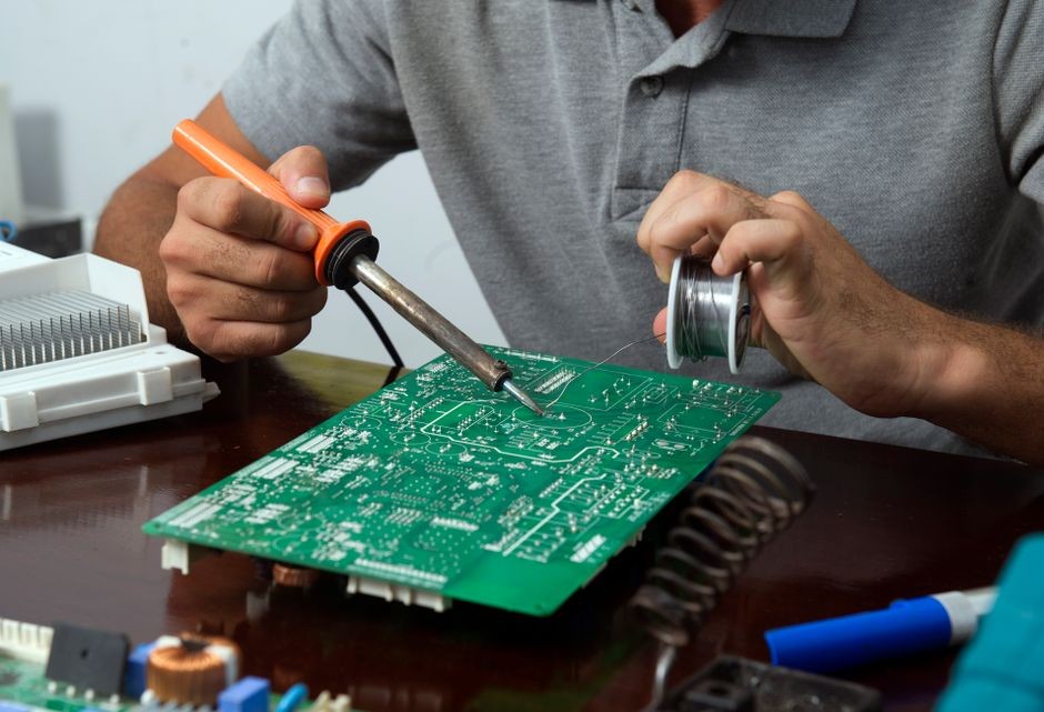 Papan sirkuit elektronik kecil sedang diperiksa dan dibersihkan di atas meja kerja