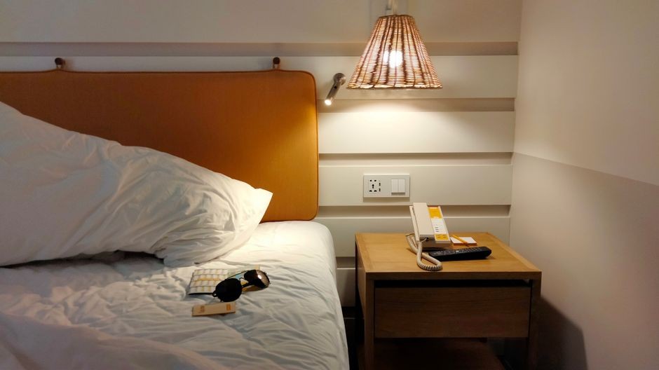 Ruang kamar yang mengundang dengan meja samping tempat tidur yang diterangi lampu, ponsel, dan kacamata hitam di atas tempat tidur.