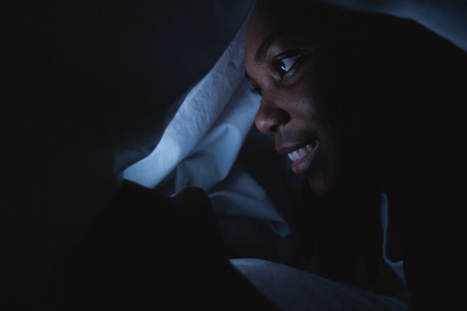 Seseorang berbaring di tempat tidur sambil menatap layar smartphone di ruangan gelap.