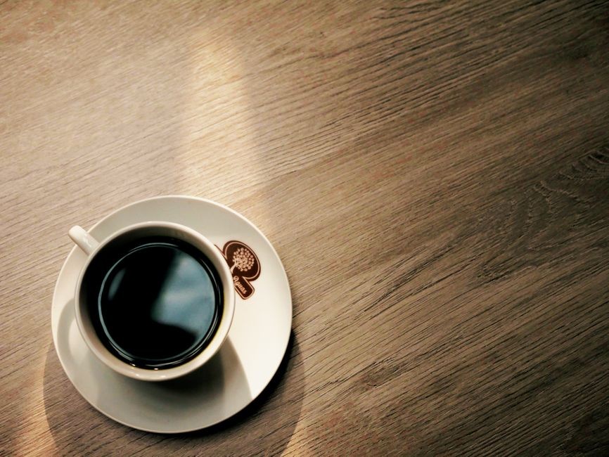 Secangkir kopi hitam mengepul di atas meja kayu pada pagi hari.