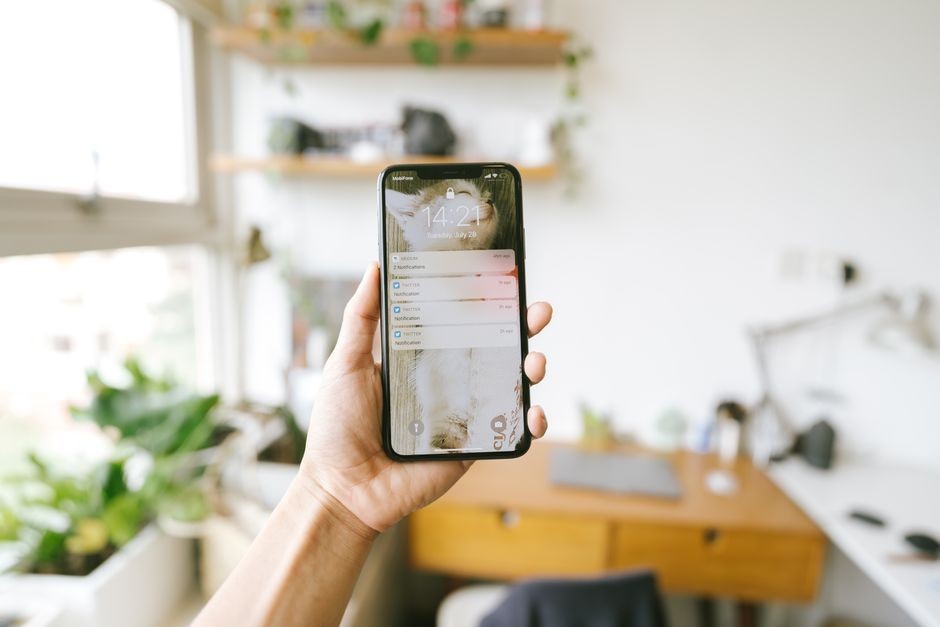 Tangan yang memegang smartphone di dalam ruangan dengan latar belakang kantor yang kabur, fokus pada notifikasi layar.