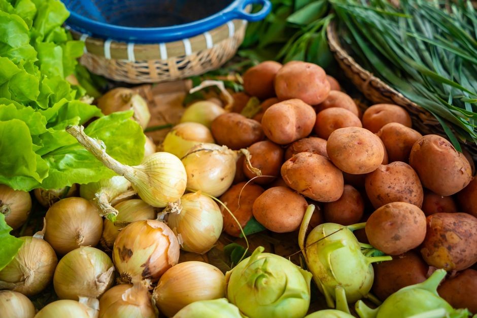 Close-up dari berbagai sayuran segar termasuk bawang, kentang, dan sayuran hijau di sebuah kios pasar petani.
