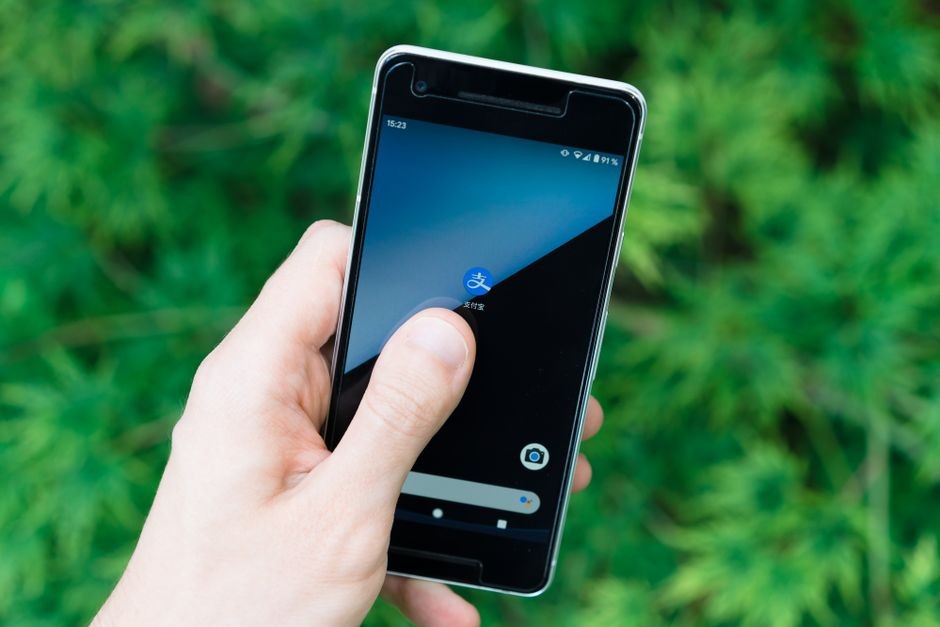 Tangan memegang smartphone dengan ikon aplikasi di layar, menunjukkan teknologi modern di luar ruangan.