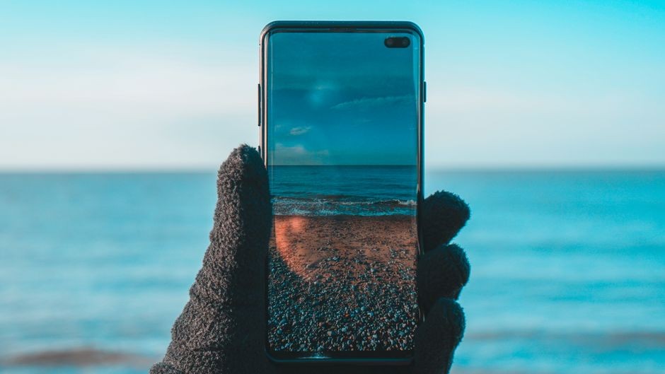 Smartphone menampilkan pemandangan pantai dan lautan di layar saat matahari terbenam.