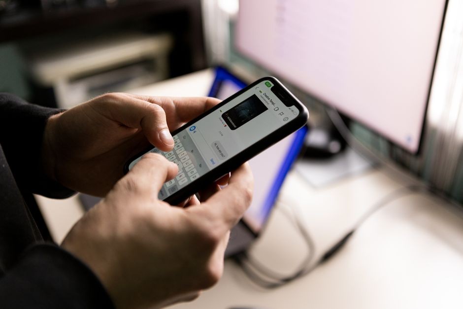 Seseorang memegang smartphone dan mengobrol di dalam ruangan dekat layar komputer.