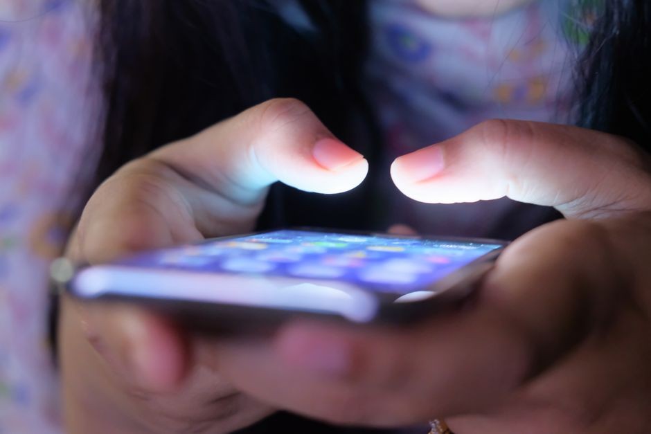 Sebuah close-up tangan yang memegang smartphone dalam cahaya rendah, menampilkan layar yang bersinar.